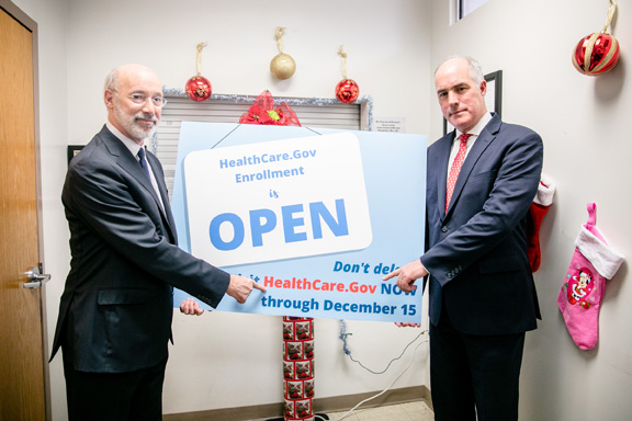Gov. Wolf and Sen. Casey holding an informational sign about Affordable Care Act enrollment and pointing at the healthcare.gov website